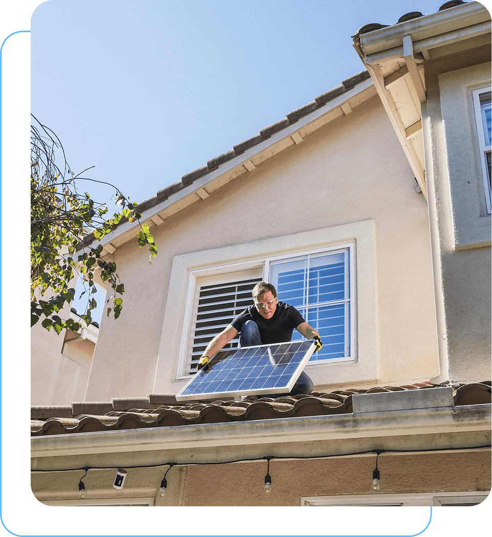 A man standing on the roof of a house holding onto a solar panel.
